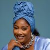 Smiling woman wearing a blue headwrap and matching satin top, with silver jewelry including a chunky necklace and ring.