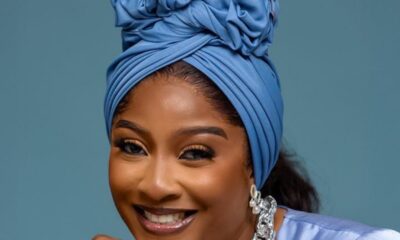 Smiling woman wearing a blue headwrap and matching satin top, with silver jewelry including a chunky necklace and ring.