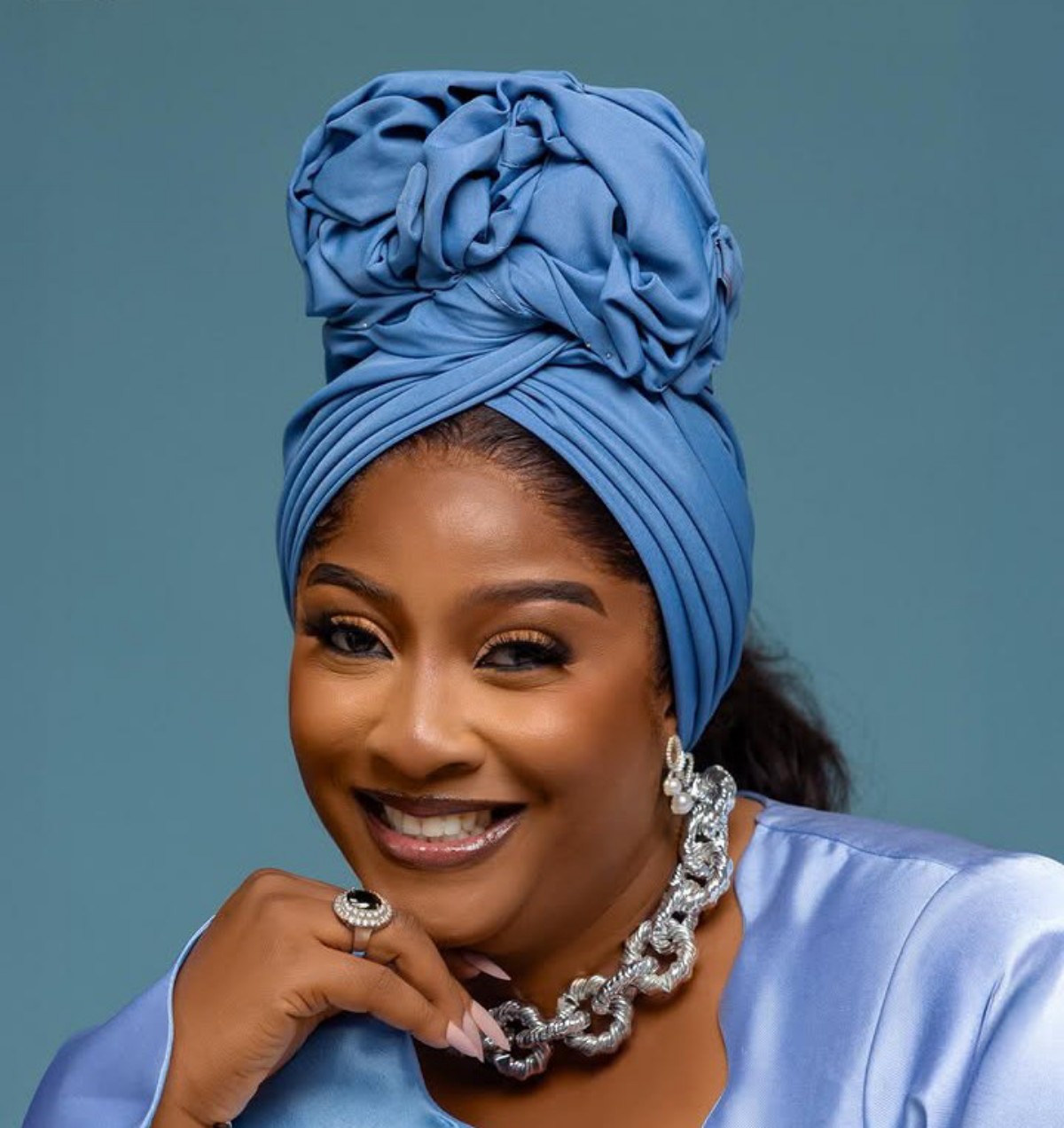 Smiling woman wearing a blue headwrap and matching satin top, with silver jewelry including a chunky necklace and ring.