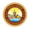 Osun State seal showing a central statue with sun rays, farm fields, a river, and gold, framed by a circular motto, ‘State of the Living Spring’.