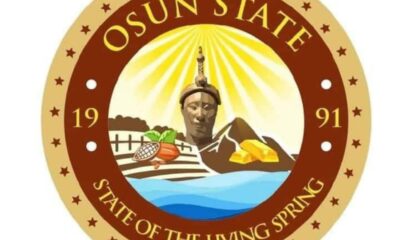 Osun State seal showing a central statue with sun rays, farm fields, a river, and gold, framed by a circular motto, ‘State of the Living Spring’.