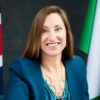 Smiling woman in teal blazer posing for a formal portrait with flags in the background.