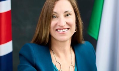 Smiling woman in teal blazer posing for a formal portrait with flags in the background.
