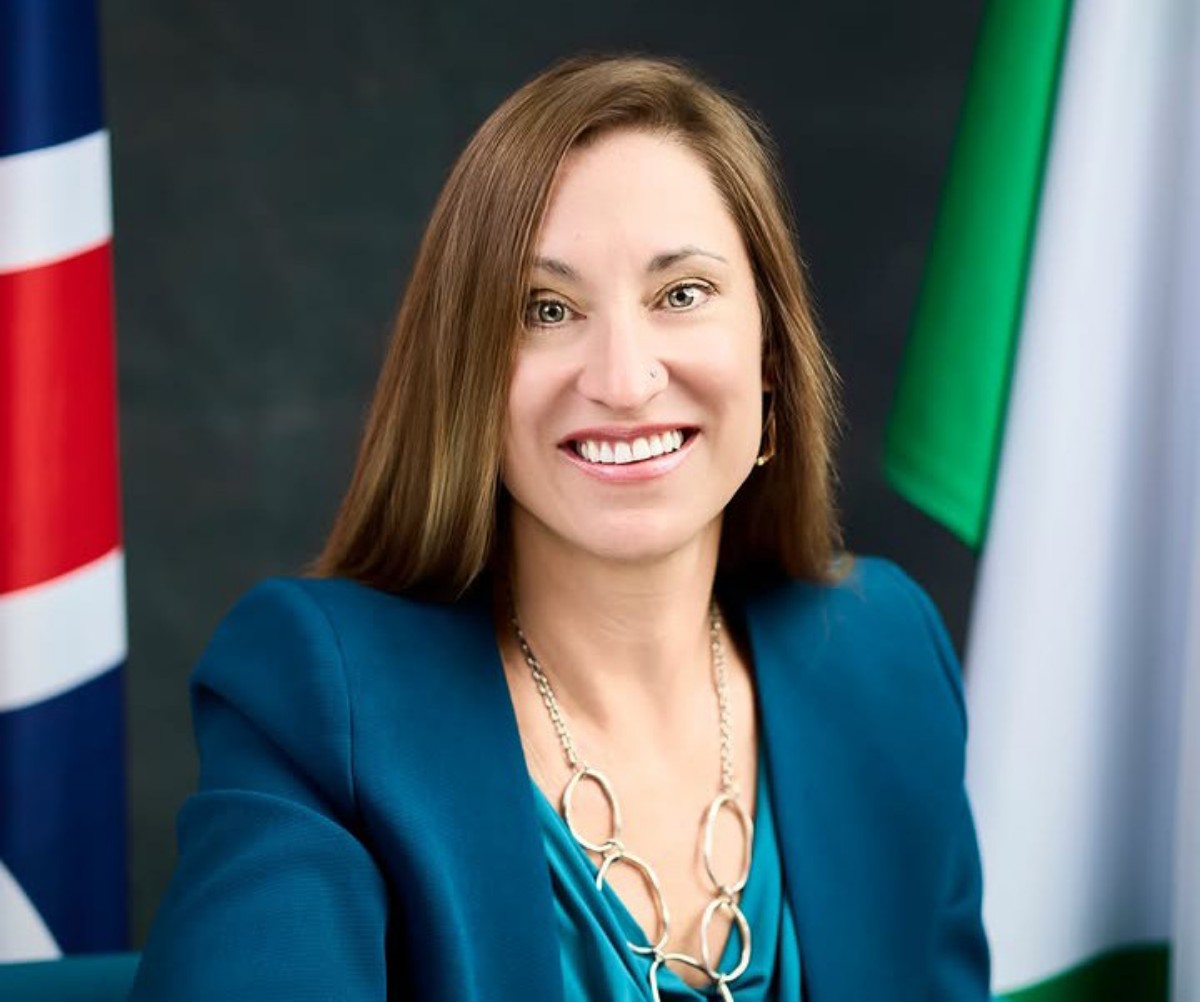 Smiling woman in teal blazer posing for a formal portrait with flags in the background.