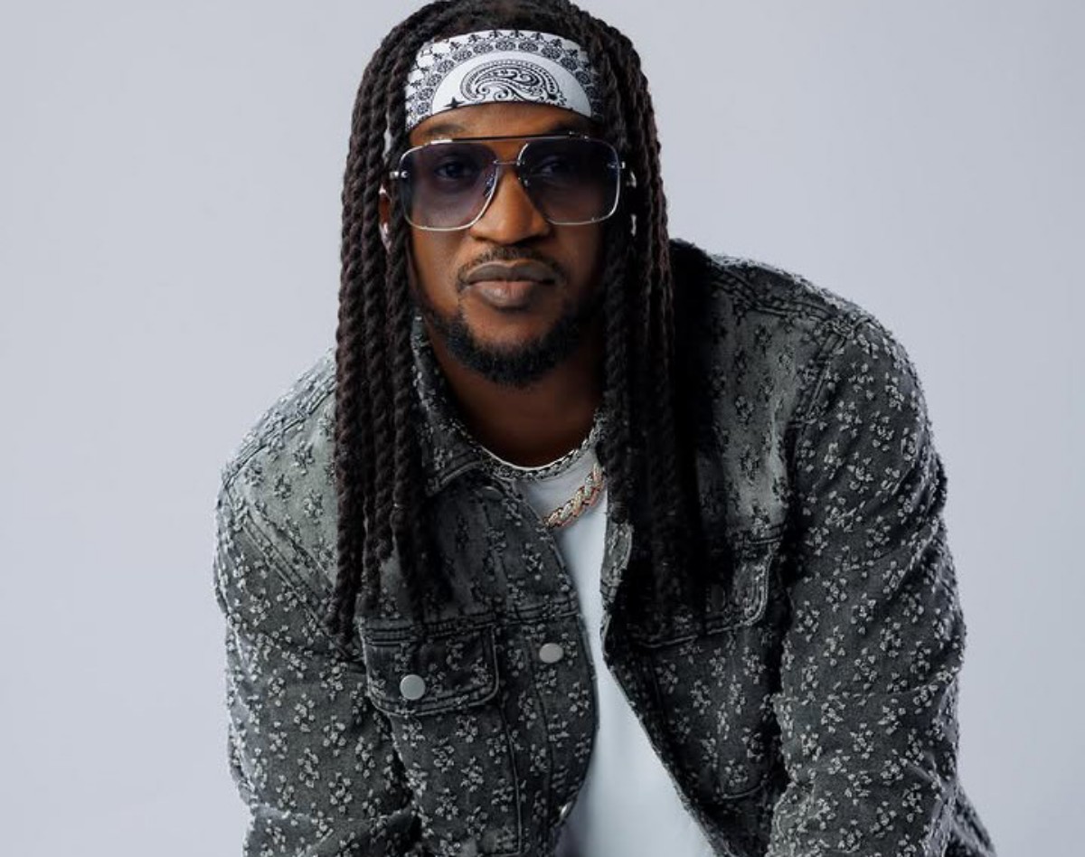 Man with long braids, bandana headband, and oversized sunglasses, wearing a floral-pattern denim jacket.