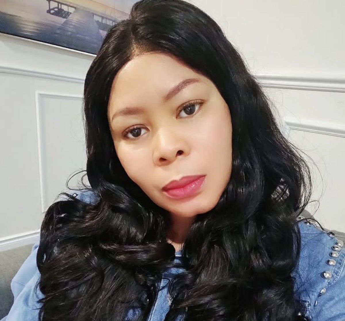 Portrait of a woman with long black hair wearing a blue denim jacket, looking at the camera indoors with a neutral expression.