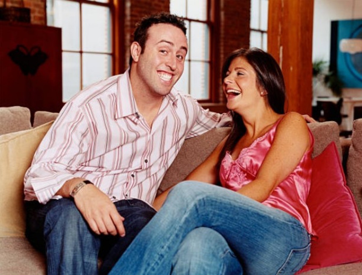 Smiling couple sitting close on a beige sofa, laughing together in a cozy living room setting.