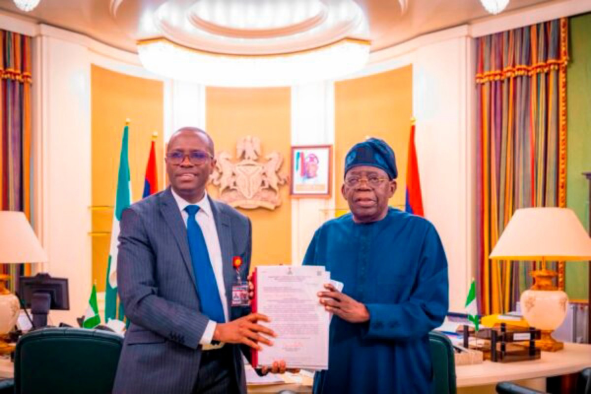 Two men in an ornate office shake hands while presenting a stack of documents; flags and crest in the background suggest an official meeting.