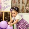 A toddler in a white top and purple skirt reaches for lilac balloons at a pastel birthday party setup with flowers in the background.