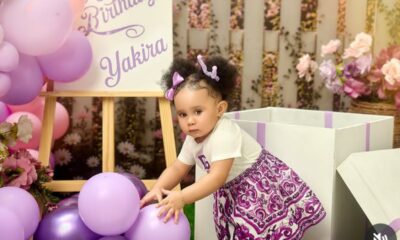 A toddler in a white top and purple skirt reaches for lilac balloons at a pastel birthday party setup with flowers in the background.
