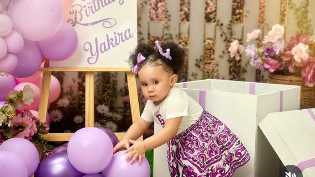 A toddler in a white top and purple skirt reaches for lilac balloons at a pastel birthday party setup with flowers in the background.