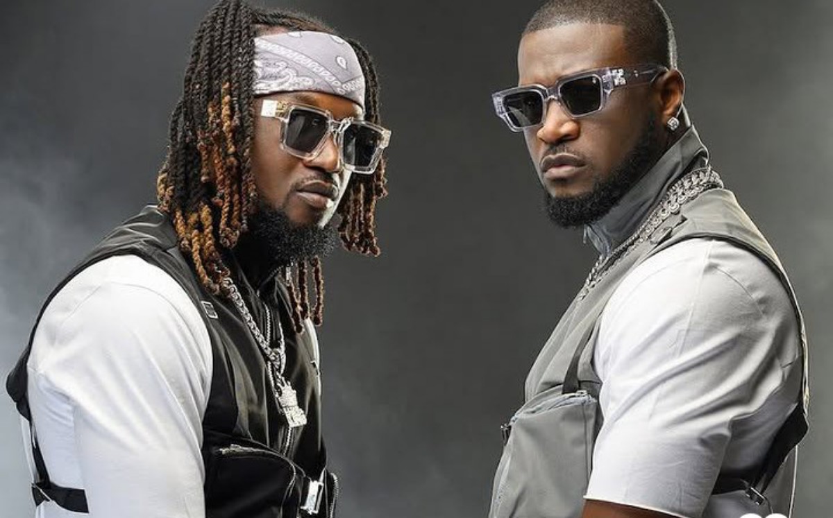 Two men pose in white shirts with gray vests, wearing sunglasses and heavy chains against a dark smoky backdrop.