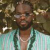 Man with braided hair and beard wearing a mint green and pink striped blazer, sunglasses, and a beaded necklace, standing in front of green leaves.