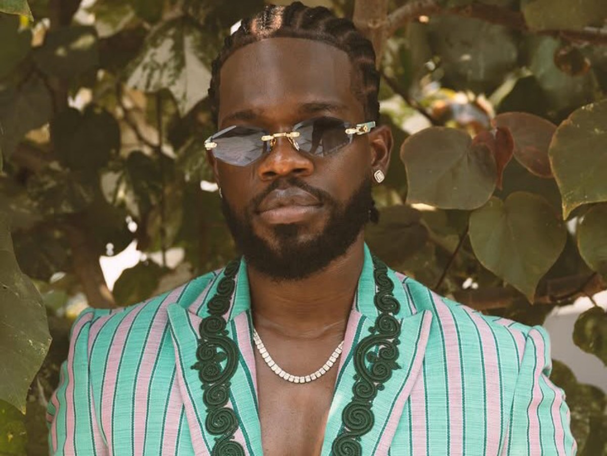 Man with braided hair and beard wearing a mint green and pink striped blazer, sunglasses, and a beaded necklace, standing in front of green leaves.