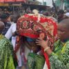 Pallbearers in green outfits carry a body wrapped in a red patterned cloth during a crowded outdoor funeral procession.