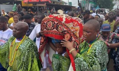 Pallbearers in green outfits carry a body wrapped in a red patterned cloth during a crowded outdoor funeral procession.