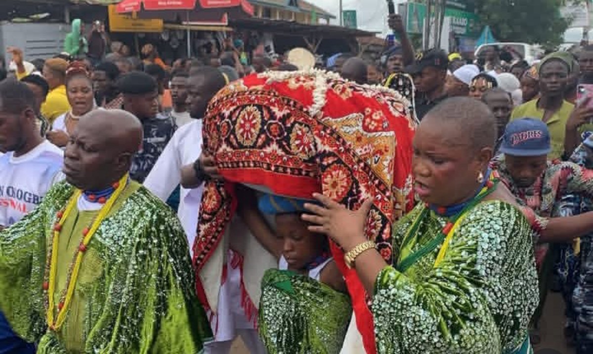 Pallbearers in green outfits carry a body wrapped in a red patterned cloth during a crowded outdoor funeral procession.