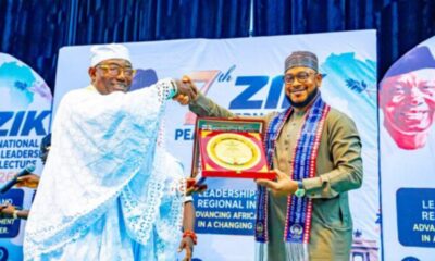 Two men on a stage shake hands as one presents a circular plaque in a red case, with blue event banners in the background.