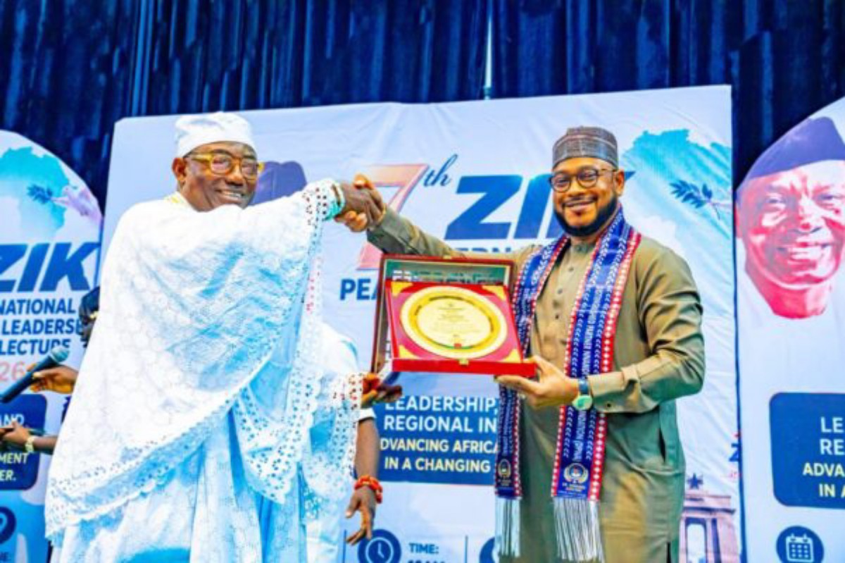 Two men on a stage shake hands as one presents a circular plaque in a red case, with blue event banners in the background.