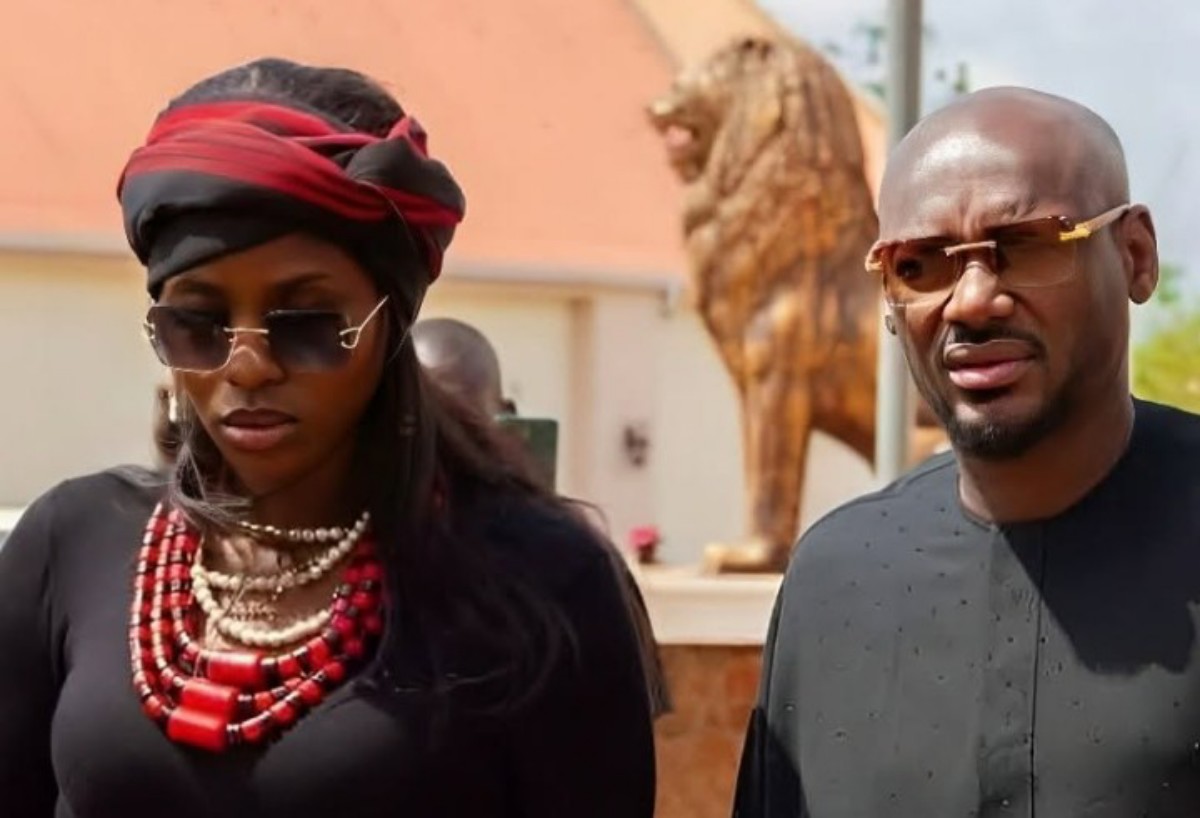 Two stylish adults wearing sunglasses outdoors; woman in a red/black headwrap with bold jewelry and the man in dark traditional attire.