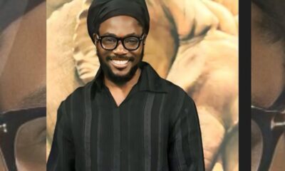 Smiling man with a black turban, black-framed glasses, and a black striped shirt posing in front of a warm, abstract backdrop.