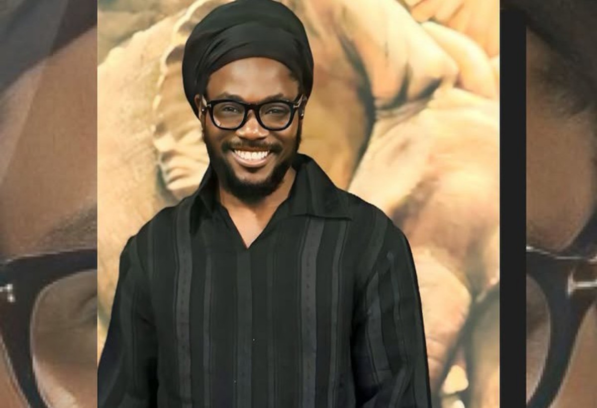 Smiling man with a black turban, black-framed glasses, and a black striped shirt posing in front of a warm, abstract backdrop.