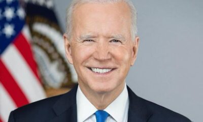 Smiling older man in a dark suit and blue tie, with an American flag in the background.