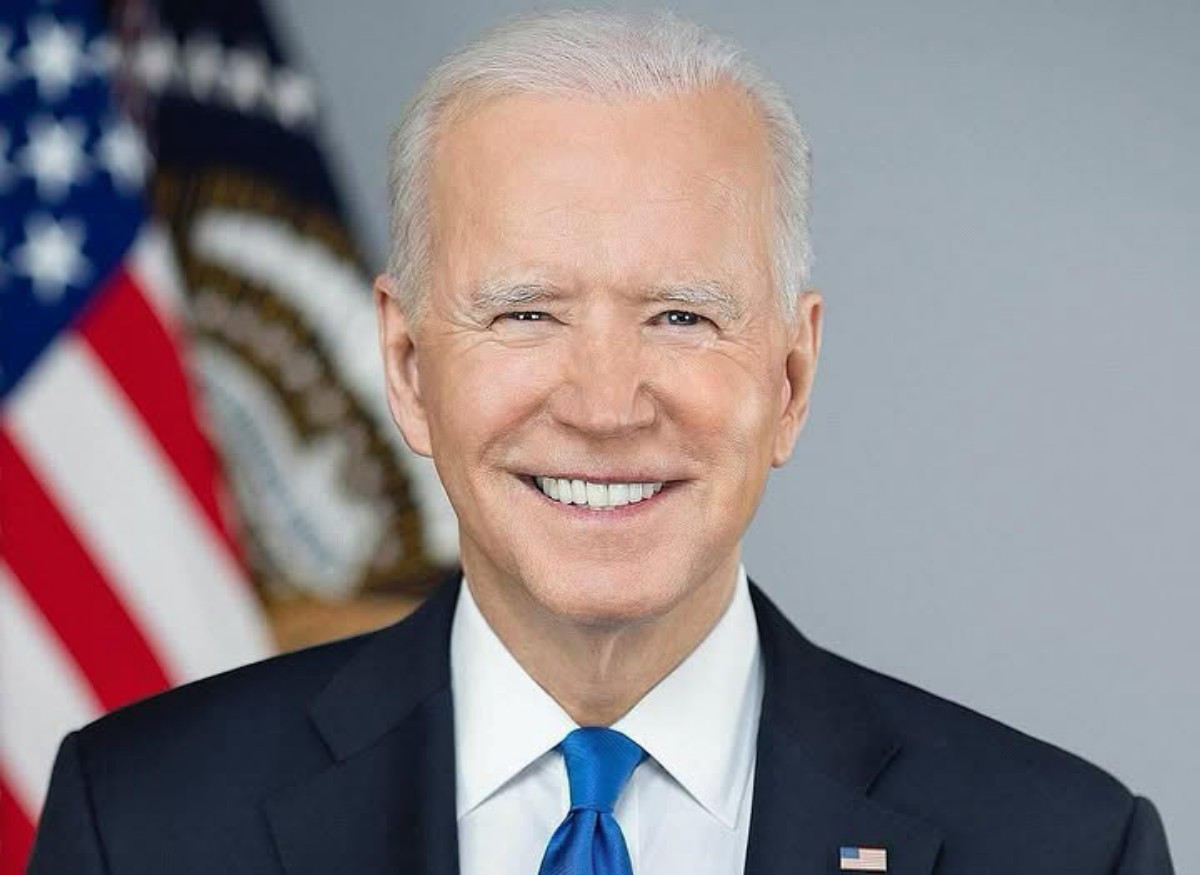Smiling older man in a dark suit and blue tie, with an American flag in the background.