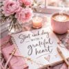 Pink floral desk scene with peonies, a lit candle, a pink mug, and a notebook reading 'Start each day with a grateful heart'.