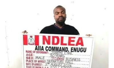 Man holding a large arrest-board with red letters that likely spell a law-enforcement agency acronym, showing fields for name, age, sex, occupation, and arrest details.