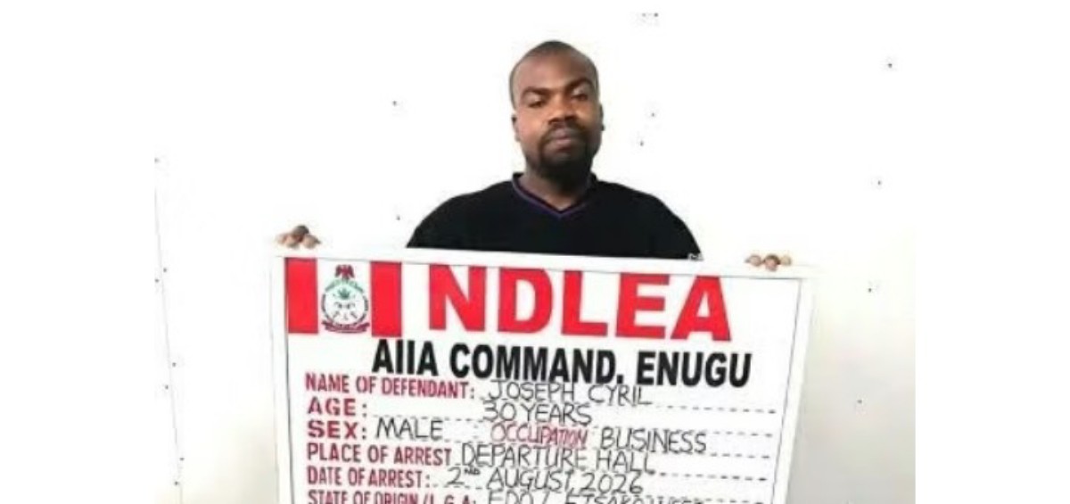 Man holding a large arrest-board with red letters that likely spell a law-enforcement agency acronym, showing fields for name, age, sex, occupation, and arrest details.