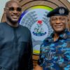 Two men smiling in front of a Nigeria Police emblem: a man in black traditional attire on the left and a police officer in blue camouflage with a beret on the right.