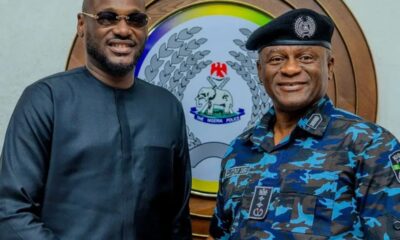 Two men smiling in front of a Nigeria Police emblem: a man in black traditional attire on the left and a police officer in blue camouflage with a beret on the right.