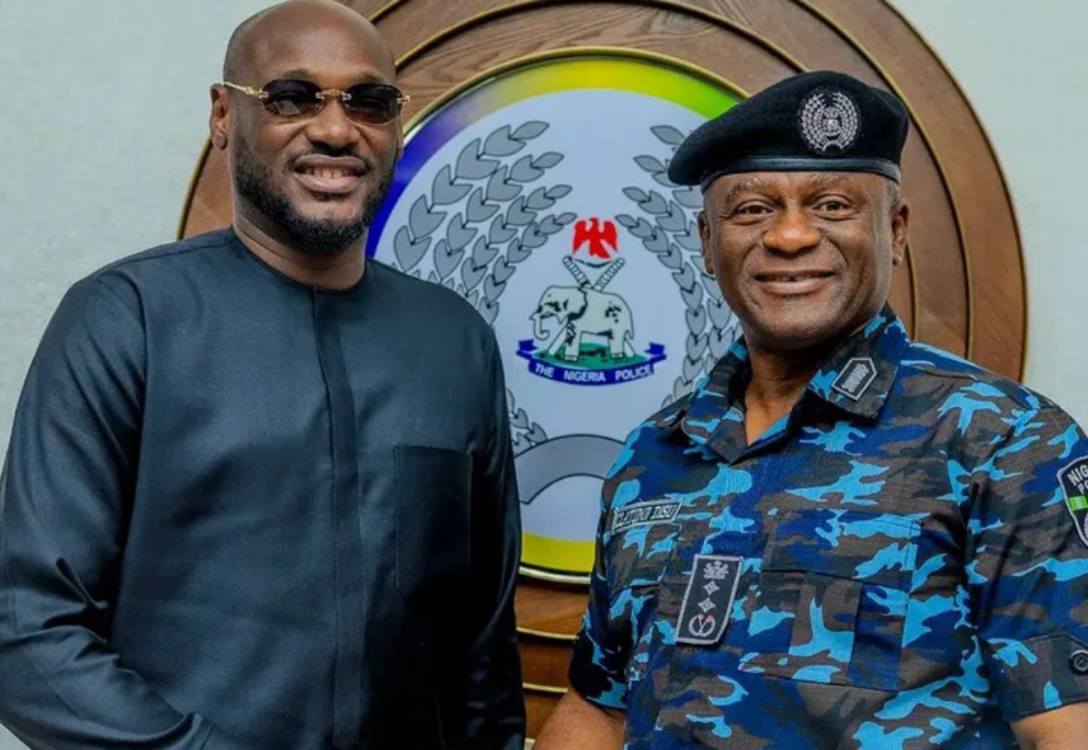 Two men smiling in front of a Nigeria Police emblem: a man in black traditional attire on the left and a police officer in blue camouflage with a beret on the right.