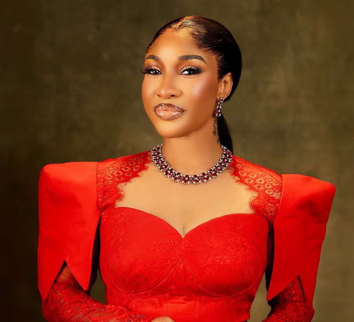 Elegant portrait of a woman in a red lace gown with a statement necklace, posed against a dark backdrop.