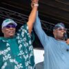 Two men on a stage raise their clasped hands in a victory gesture; one wears a green traditional agbada and cap, the other a blue outfit with a headscarf and sunglasses, holding a microphone.