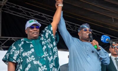 Two men on a stage raise their clasped hands in a victory gesture; one wears a green traditional agbada and cap, the other a blue outfit with a headscarf and sunglasses, holding a microphone.