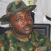 Nigerian soldier in camouflage uniform and cap, seated indoors, looking to the side.