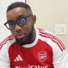 Man with short hair and beard wearing glasses in an Arsenal jersey indoors in a kitchen setting.