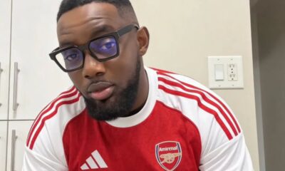 Man with short hair and beard wearing glasses in an Arsenal jersey indoors in a kitchen setting.
