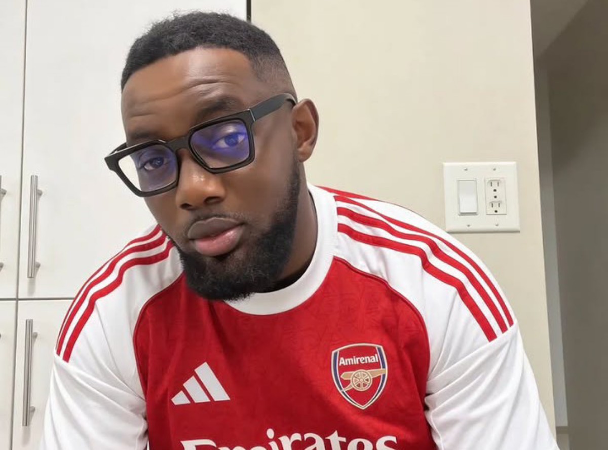 Man with short hair and beard wearing glasses in an Arsenal jersey indoors in a kitchen setting.