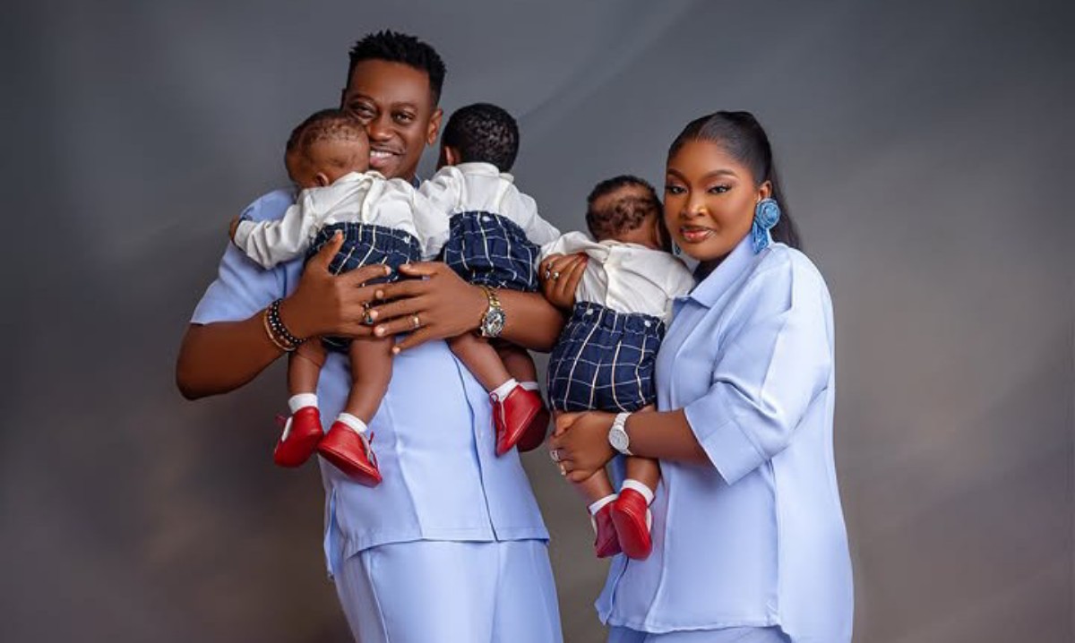 Couple in matching blue outfits cradle four babies in coordinated white tops, navy shorts, and red shoes for a family portrait.