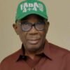 Portrait of a man wearing a green cap with white lettering, glasses, and a brown shirt.