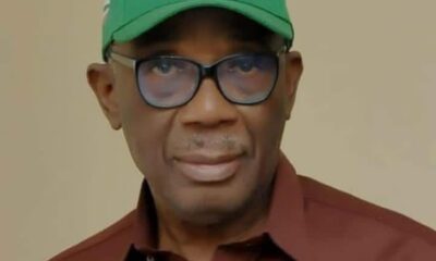 Portrait of a man wearing a green cap with white lettering, glasses, and a brown shirt.