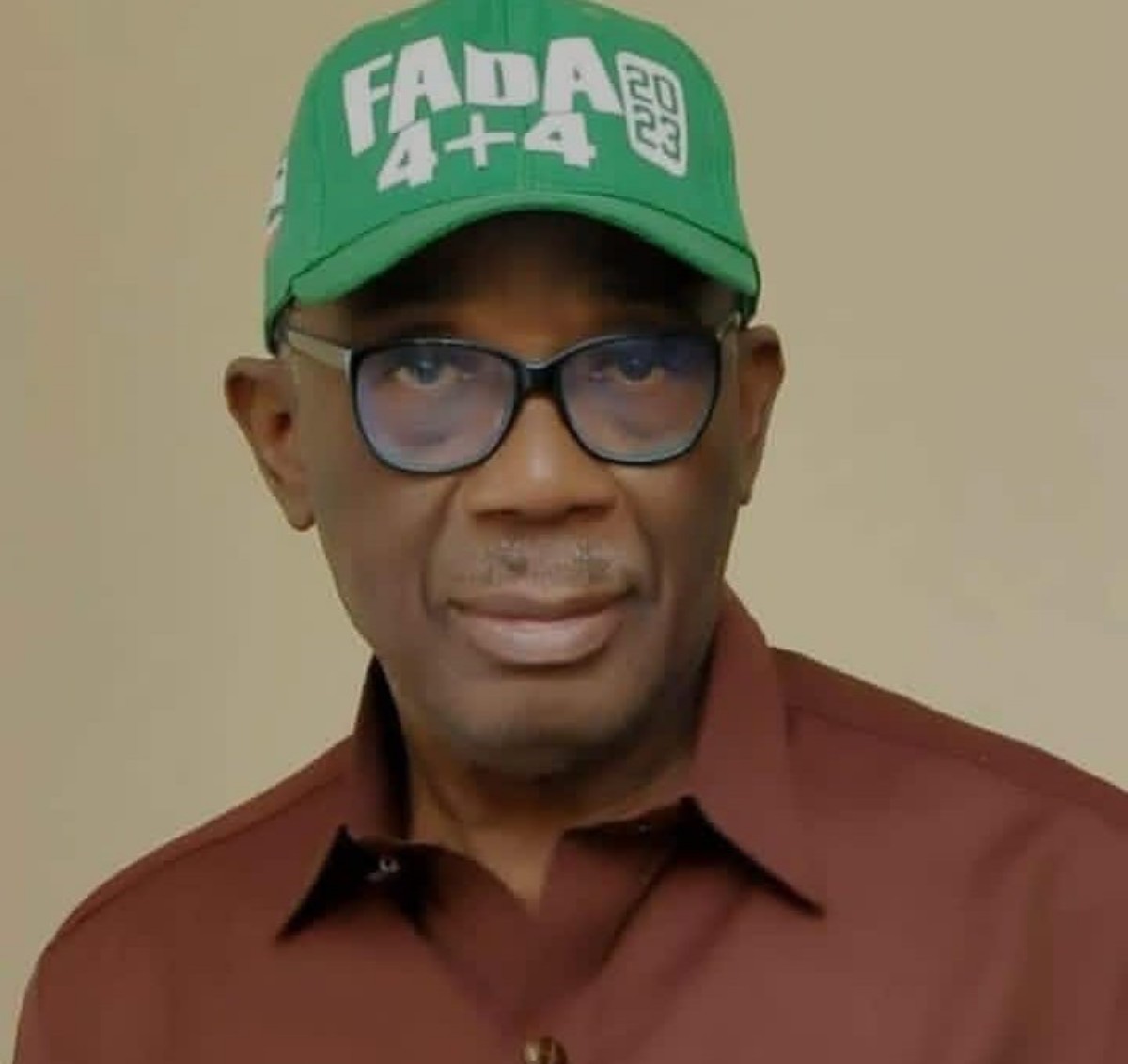 Portrait of a man wearing a green cap with white lettering, glasses, and a brown shirt.