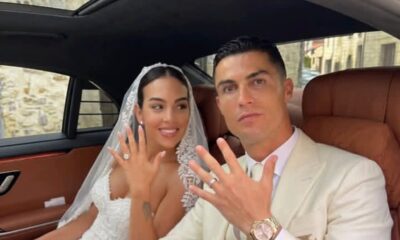 Bride and groom showing their wedding rings while sitting in a car, smiling at the camera.