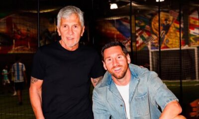 Two men posing together indoors at a sports facility, one in a black t-shirt and the other in a denim jacket, smiling at the camera.