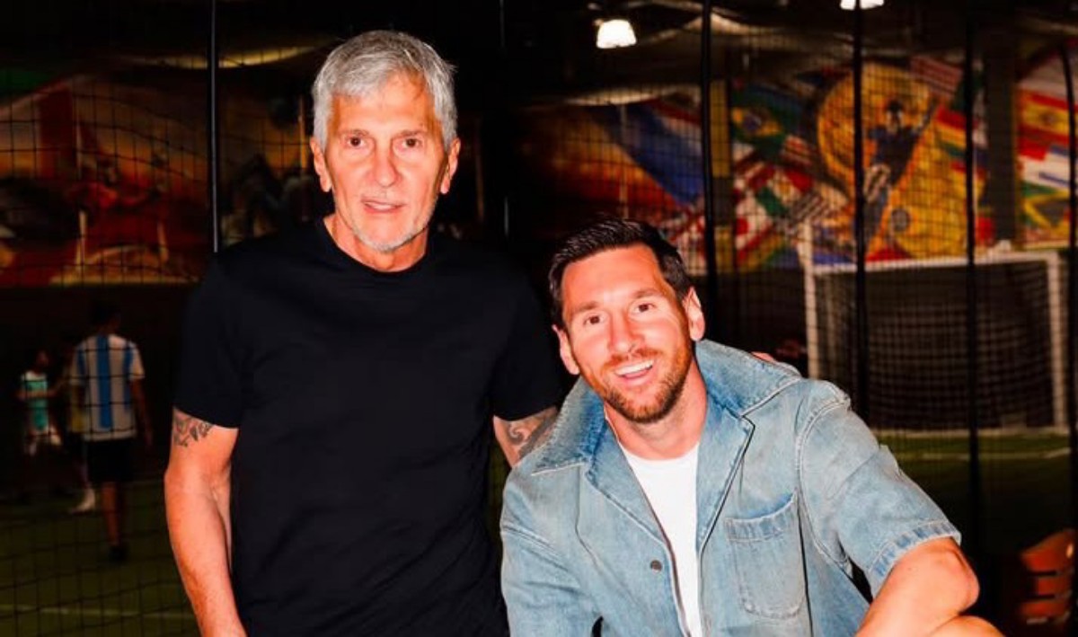Two men posing together indoors at a sports facility, one in a black t-shirt and the other in a denim jacket, smiling at the camera.