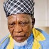 Portrait of an elderly man with a black-and-white plaid head wrap, wearing blue and yellow traditional clothing, looking at the camera.
