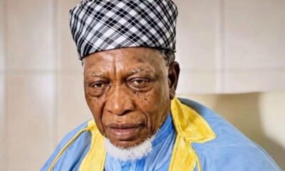 Portrait of an elderly man with a black-and-white plaid head wrap, wearing blue and yellow traditional clothing, looking at the camera.